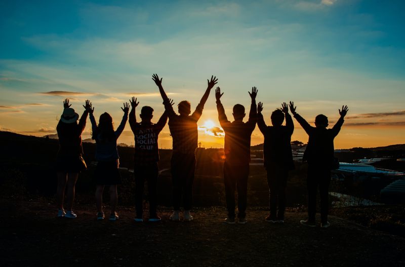 People Raise Hands Up Against Evening Sun Background.